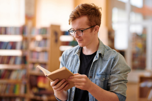 Młody mężczyzna w okularach stoi bokiem i trzyma w ręku książkę. Czyta. W tle znajdują się regały z książkami
