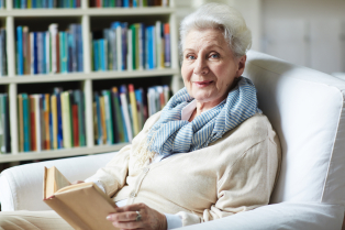 Na zdjęciu starsza kobieta siedzi w białym fotelu, trzymając w rękach książkę, jednocześnie uśmiecha się do obiektywu. W tle widać regał z książkami. 