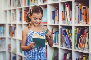 Na zdjęciu dziewczynka stoi przy regałach bibliotecznych pełnych książek i czyta książkę, którą trzyma w rękach.